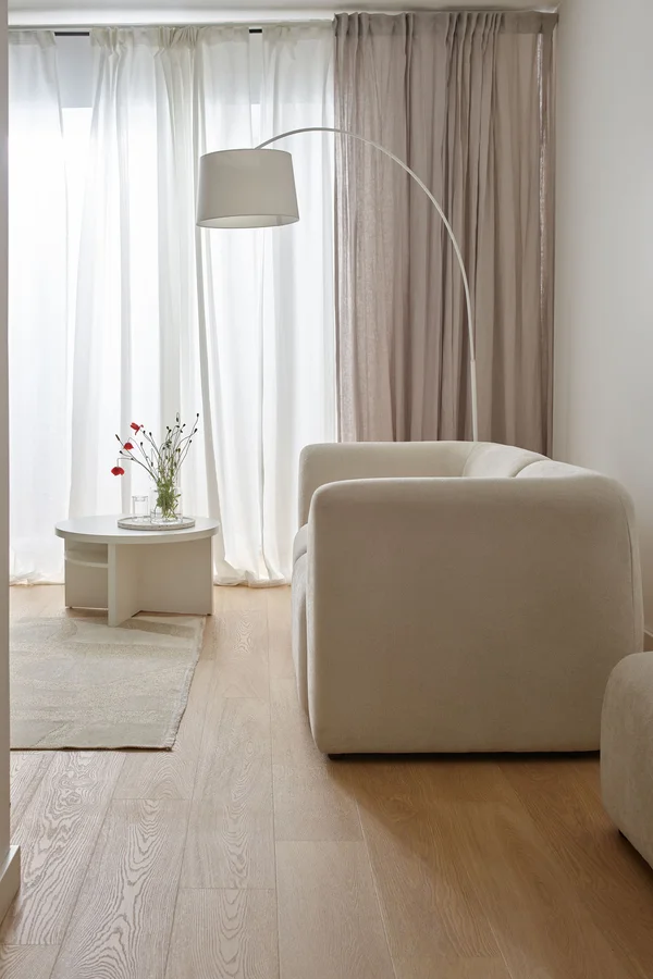 Biała sofa ustawiona obok dużych okien z jasnymi zasłonami, które wpuszczają naturalne światło i wizualnie podnoszą sufit.